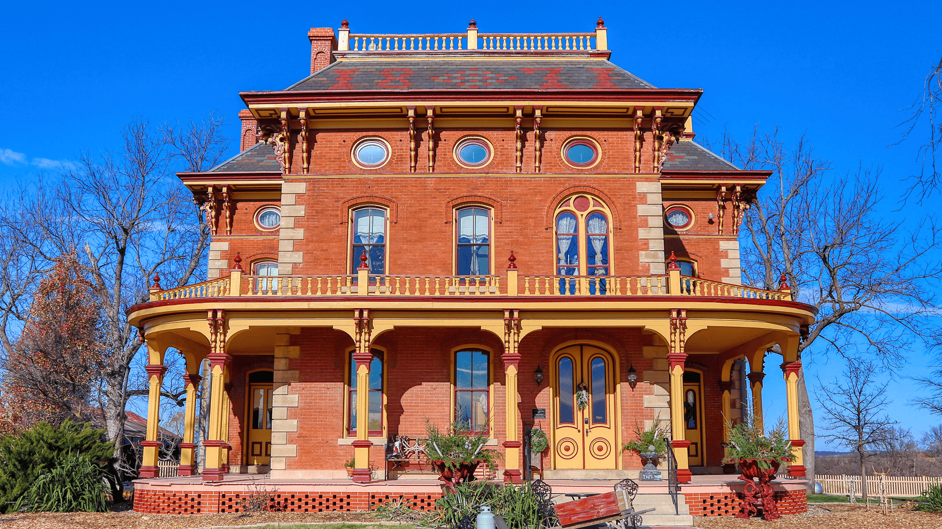 A historic red brick house with yellow trim and a wraparound porch under a clear blue sky.