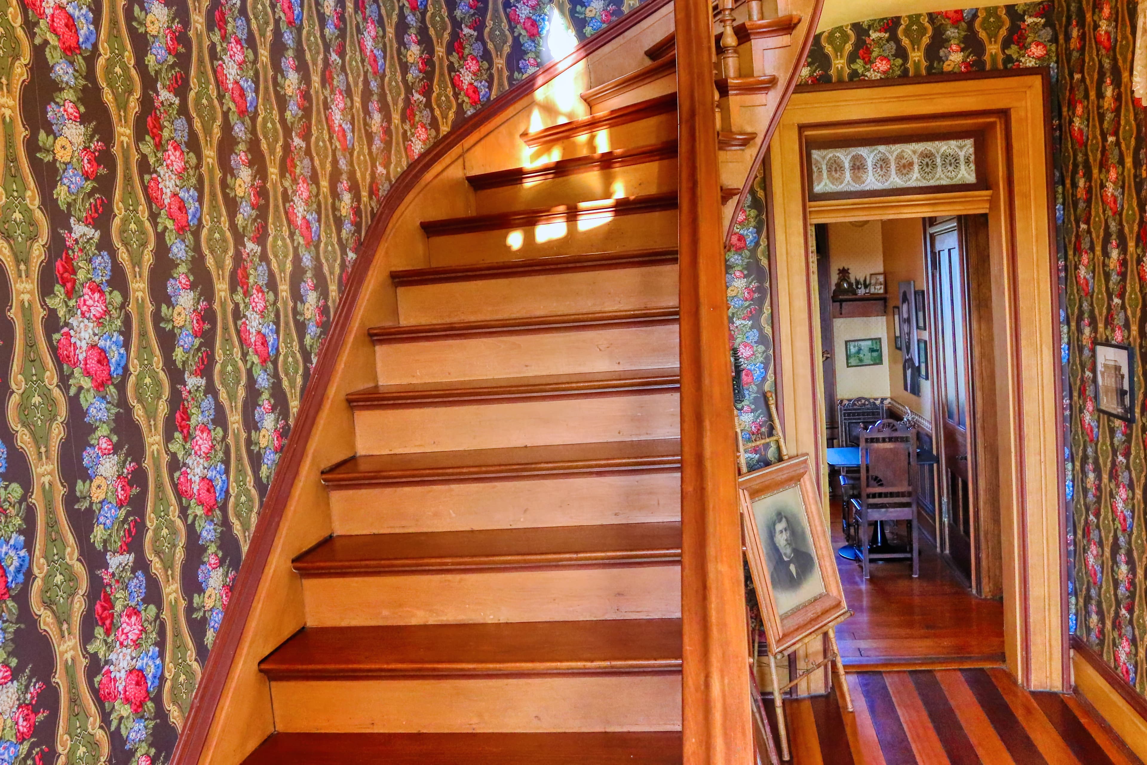 A spiral staircase leads upstairs, flanked by floral wallpaper and an open doorway to a cozy room.